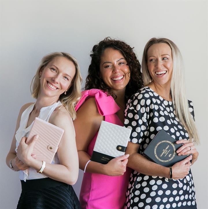 A support network of women stand together smiling, holding LH Agenda journals.