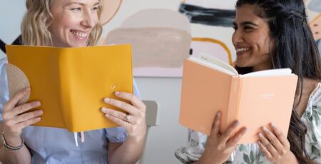Two women hold up their LH Agenda planners from behind a desk and smile at eachother.