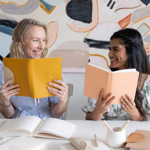 Two women hold up their LH Agenda planners from behind a desk and smile at eachother.