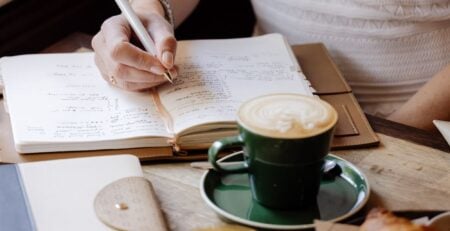 A woman writes in her planner at a cafe table with a coffee, croissant and other stationery items.