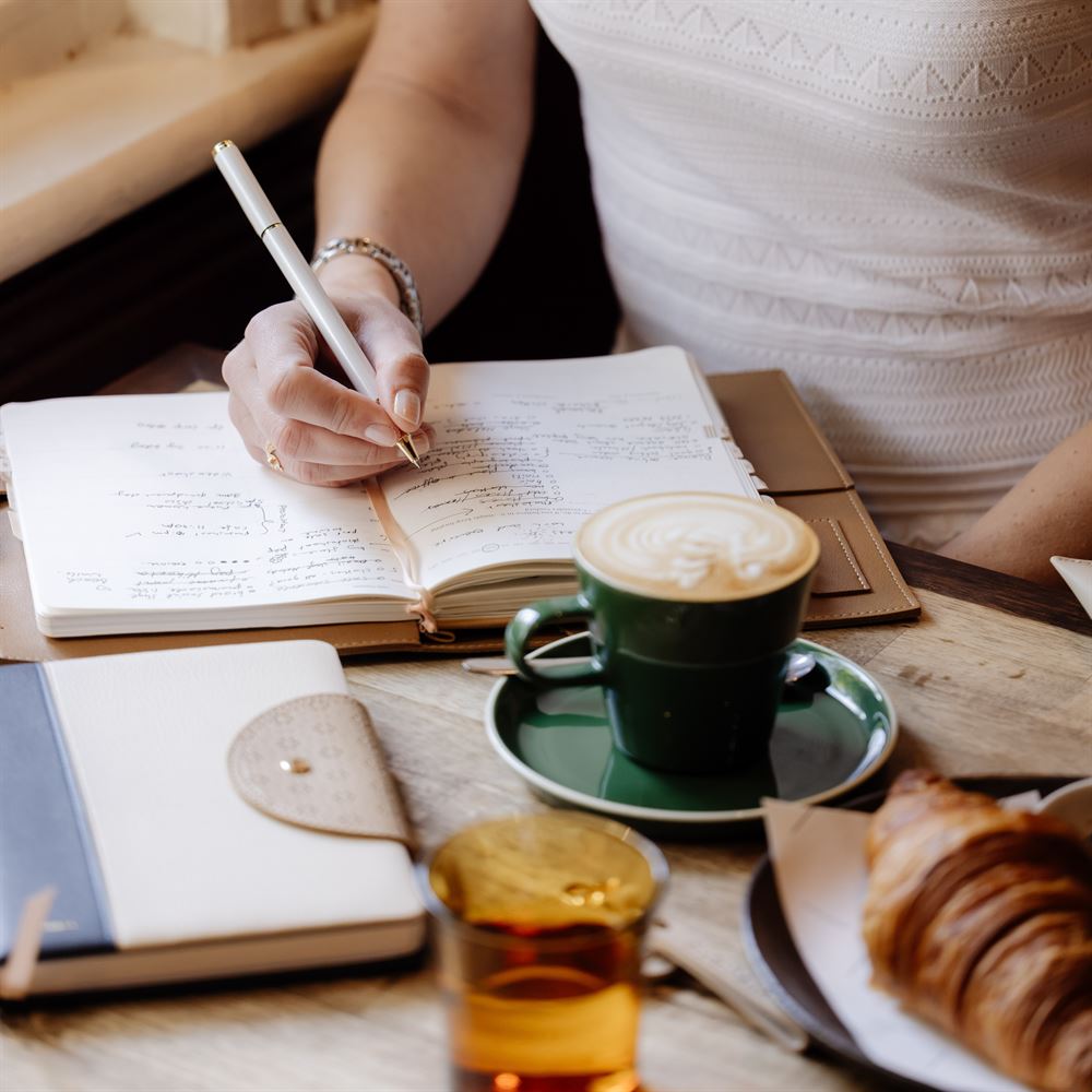 A woman writes in her planner at a cafe table with a coffee, croissant and other stationery items.