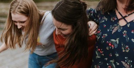 Three female friends have their arms around eachother, laughing, smiling and living with intention.