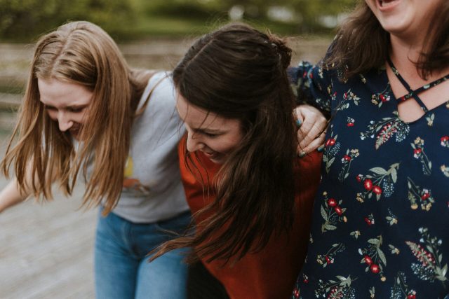 Three female friends have their arms around eachother, laughing, smiling and living with intention.